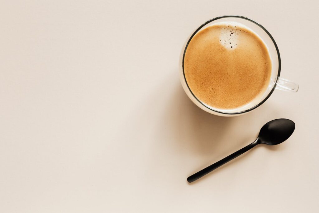 glass of hot cappuccino near spoon on beige background